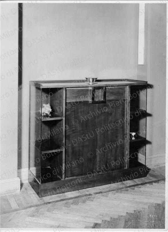 Veduta di "Credenza di salotto", positivo, bianco e nero, mm 227x168. Timbri ad inchiostro sul verso: "Piero Bottoni" e "La proprietà di questo modello è protetta a norma dell'art. 1 e seg. Del R.D.del 7 nov. 1925 n. 1950 sul diritto d'autore". Annotazione manoscritta sul verso: "Credenza di salotto in legno di pero lucidato verde e nero. Cassetti ruotanti (chiusi) spalle in cristallo, maniglie in metallo nichelato. Asse allungabile per servizio da tavola".
