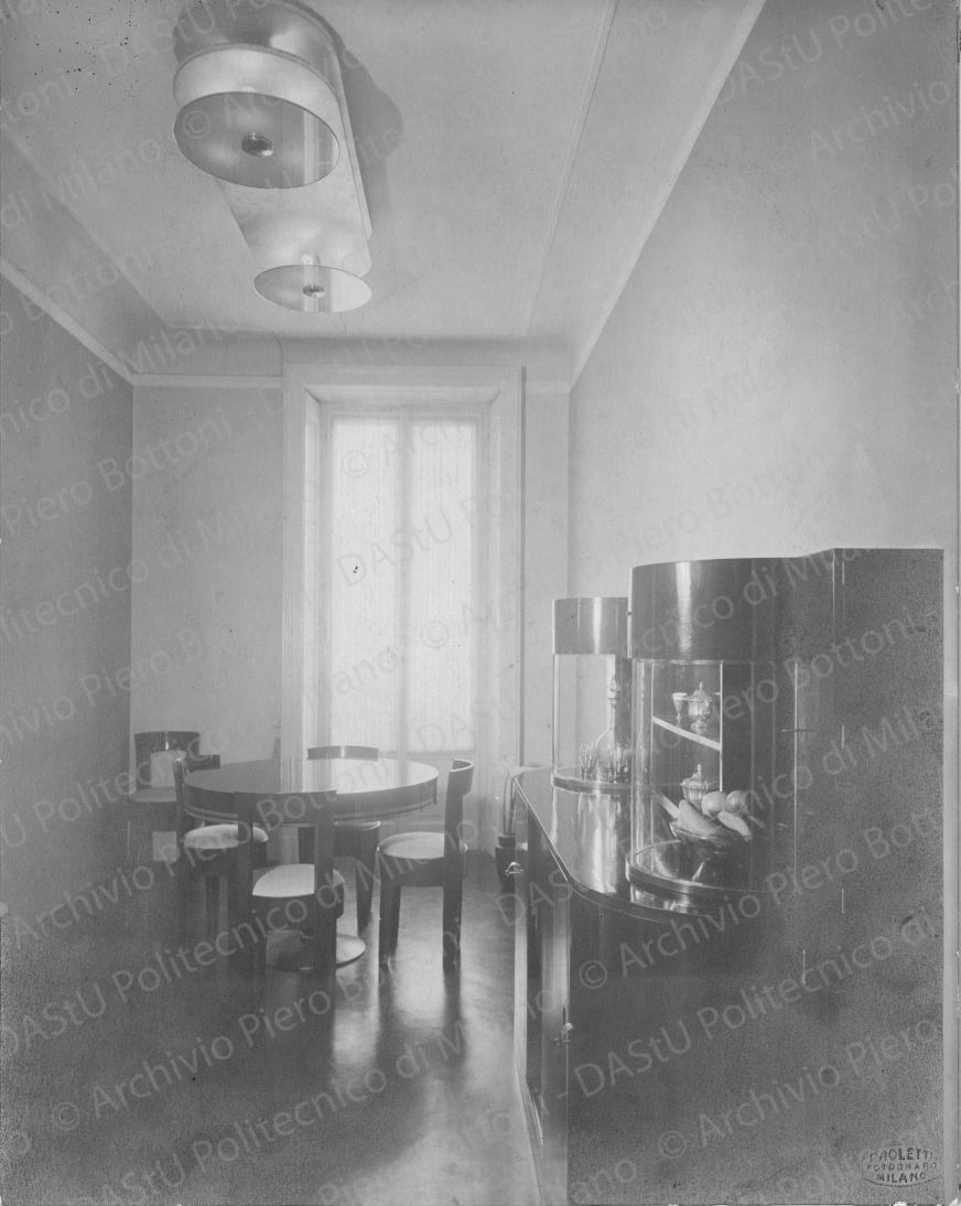 Veduta della camera da pranzo. Didascalia dattiloscritta: "Camera da pranzo nell'appartamento ing.Ma Milano. Pavimento in linoleum nero; pareti a due colori: gialla e grigio azzurro; tavolo e seggiole in legno di pero lucidato verde oliva scurissimo e nero; finiture in metallo bianco; buffet in pero lucidato come sopra, con sportelli scorrevoli, ripiani, ripostigli, cristalliere a vetri curvi scorrevoli. Apparecchio di illuminazione a luce diretta e indiretta bianco e dischi di specchio trasparente.". Timbro a secco "Paoletti fotografo Milano". Positivo, bianco e nero, 280x220 mm,  album giallo n.8, pag.62.