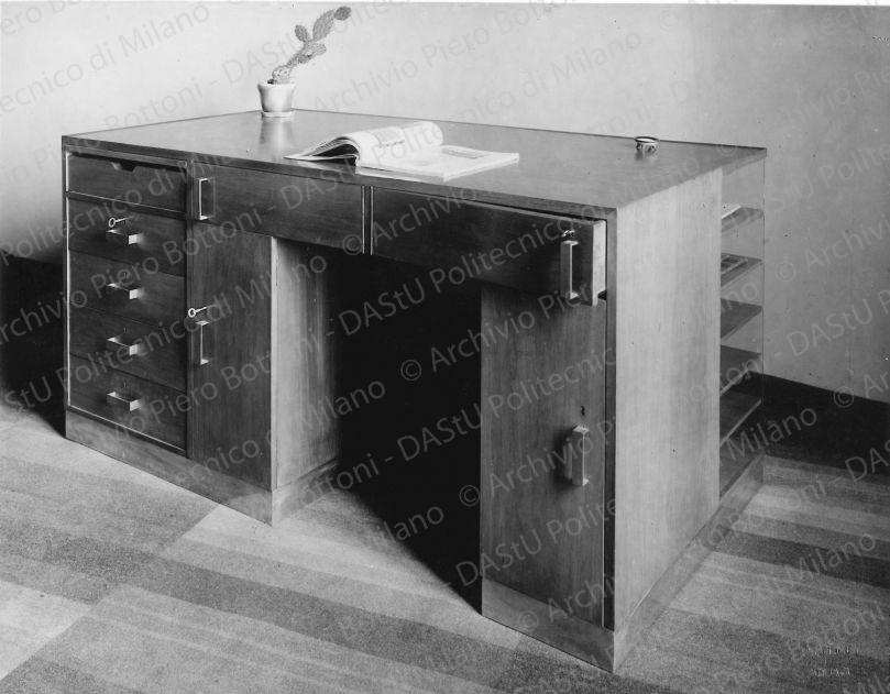Veduta di scrivania. Didascalia dattiloscritta: "(Appartam. Ing.M. in Milano) Scrivania in noce naturale chiaro. Zoccolo in metallo bianco; piano superiore in linoleum; scomparti, armadietti, cassetti vari; ripiani con spalla in metallo; cassetti ruotanti che permettono di essere aperti senza spostamento della persona che siede davanti alla scrivania". Timbro a secco "Paoletti fotografo Milano". Positivo, bianco e nero, 160x206 mm, album giallo n.8, pag.66.