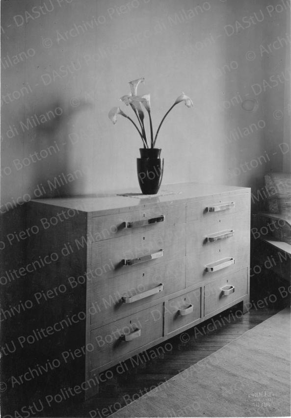 Veduta di cassettiera. Didascalia dattiloscritta: "Appartamento Ing. Minerbi - Milano. Canterale nella camera da letto; in acero grigio fr*** e piano in rabano bianco. Maniglie in metallo cromate. I cassetti a basso sono ruotanti. Vaso tornito in serpentino.". Timbro a secco "Paoletti fotografo Milano". Positivo, bianco e nero, 217x150 mm, album giallo n.8, pag.70.