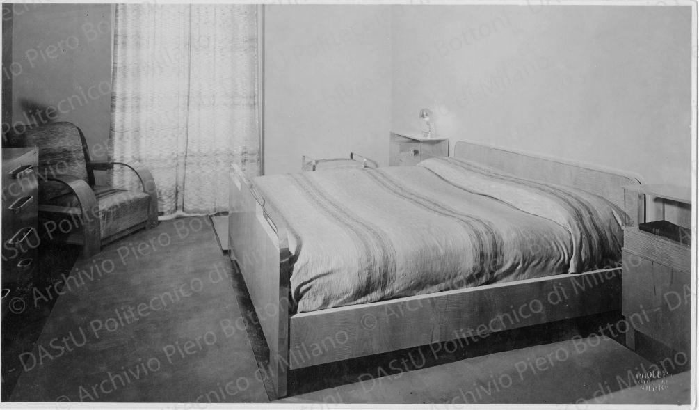 Veduta della camera da letto, positivo, bianco e nero, mm 166x285. Su le fotografie timbro ad inchiostro sul verso: "Piero Bottoni". Su una fotografia timbro ad inchiostro sul verso: "La proprietà di questo modello è protetta a norma dell'art. 1 e seg. Del R. D. L. del 7 nov. 1925 n. 1950 sul diritto d'autore" e annotazione dattiloscritta sul verso: "Appartamento Ing. Minerbi - Milano. Camera da letto in acero grigio fris? e finiture in metallo cromato, poltrona, sgabello, comodini, letto".