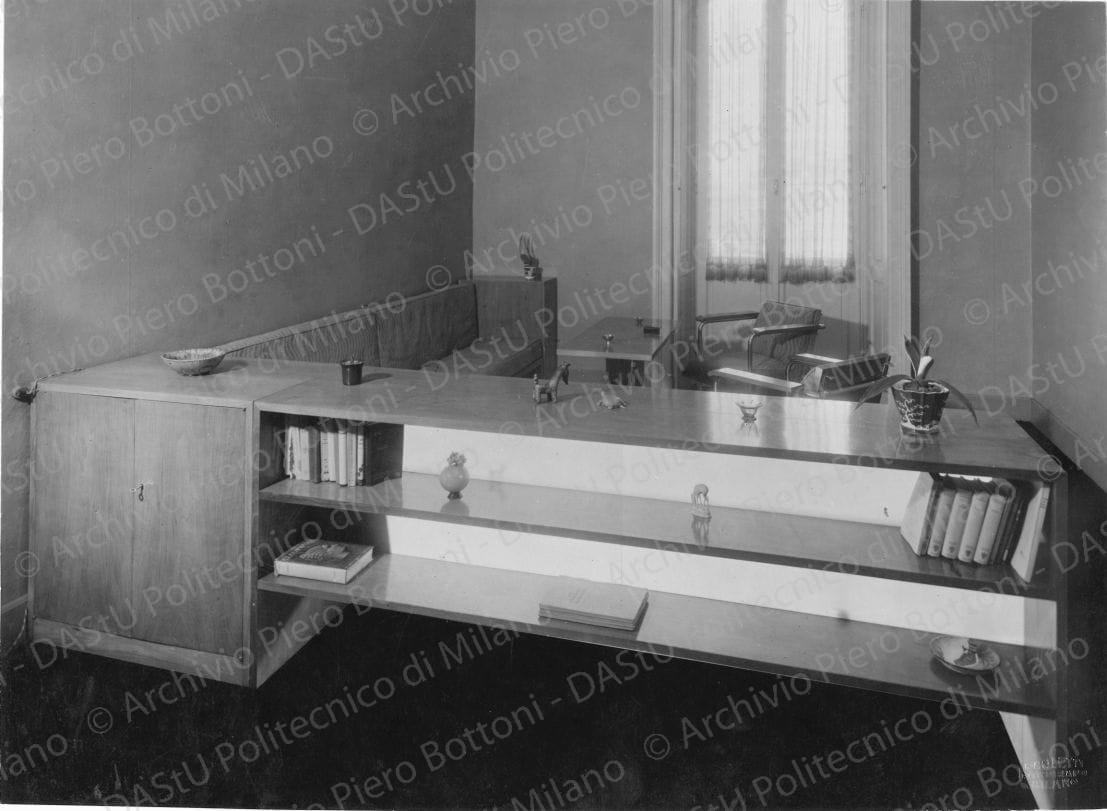 Veduta dello studio. Didascalia manoscritta: "Casa Franceschini, Milano. Locale di soggiorno e di lavoro. In primo piano scaffale libreria a doppia pancia con anta divisoria in vetro placcato paglierino. A sinistra armadietto da lavoro. Nello sfondo divano, tavolino, poltrone in tubo di acciaio, mobiletto per lavoro. A terra linoleum rosso arancione. Legno in noce chiaro. Tappezzerie Terra di Siena e bianco-sporco". Timbro a secco "Paoletti fotografo Milano". Positivo, bianco e nero, 205x280 mm, album giallo n.8, pag.50.