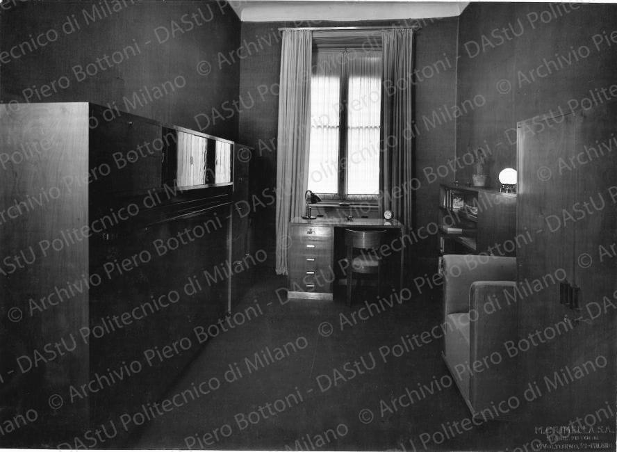Veduta della camera-studio. Didascalia manoscritta: "Casa Minetti. Camera - studio in Milano. A sinistra letto ribaltabile con vetrina per bigiotterie (?) e cassetti. Dietro libreria chiusa. Al centro scrivania. A destra libreria a giorno e **** piani mobili per abiti". Timbro a secco del fotografo "M. Crimella S.A. Stabil. Fotogr. V. Volturno 39 Milano". Positivo, bianco e nero, 164x227 mm, album giallo n.8,pag.84.
