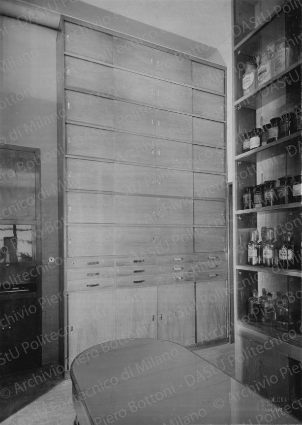 Veduta del banco di vendita e degli armadi dei medicinali. Timbro a secco "Paoletti fotografo Milano". Positivo, bianco e nero, 219x156 mm, album giallo n.6, pag.9.