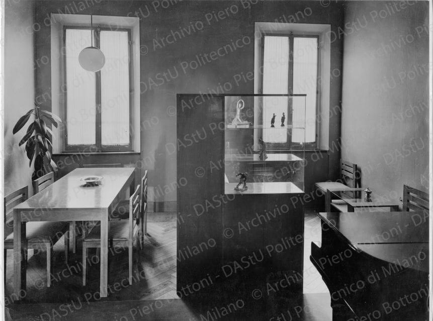 Veduta della "Sala da pranzo e salottino da ricevimento e da musica", positivo, bianco e nero, mm221x290. Timbri ad inchiostro sul verso: "Piero Bottoni", "Da restituire" e "La proprietà di questo modello è protetta a norma dell'art. 1 e seg. Del R. D. L. del 7 nov. 1925 n. 1950 sul diritto d'autore". Annotazione dattiloscritta su carta velina incollata sul verso della fotografia: "Casa Avv. C. in Ferrara. Sala da pranzo e salottino da ricevimento e da musica. Il locale è diviso in due parti da un complesso di mobili che taglia la stanza creando i due spazi destinati a due specifiche distinte funzioni: di tale complesso di mobili alcuni sono accessibili da sinistra (armadi e ripiani per servizi di tavola) altri in testa (vetrina per ceramiche e vetri d'arte e armadio per libri di musica) altri infine sono utilizzabili dalla parte del salottino (libreria e divano); tutti i mobili sono in legno di noce chiaro; il piano in gomma verde chiaro; le stoffe grigio e grigio verde a strisce sfumate; la vetrina con piani e fianchi in cristallo ha i ripiani illuminabili di costa illuminanti gli oggetti esposti. Pareti con tappezzerie in carte naturali su toni terra Siena; in primo piano (a terra) striscia di tappeto verde chiaro".