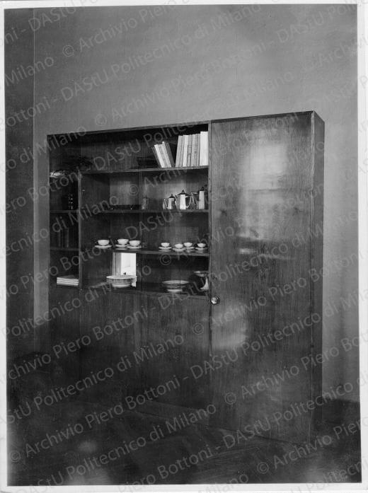 Veduta del "Mobile grande della sala da pranzo", positivo, bianco e nero, mm 236x176. Timbro ad inchiostro sul verso: "Piero Bottoni". Annotazione dattiloscritta su carta velina incollata sul verso della fotografia: "Mobile grande della sala da pranzo per servizi di tavola e libri; superiormente vani a giorno al centro scomparti con cristalli scorrevoli e passapiatti; in basso ante in legno e a destra doppia porta verso la cucina".