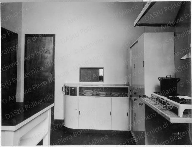 Veduta della cucina, positivo, bianco e nero, mm 161x216. Timbri ad inchiostro sul verso: "Piero Bottoni" e "Da restituire". Annotazione dattiloscritta su carta velina incollata sul verso della fotografia: "Cucina in legno laccato bianco alla nitro. Guarnizioni in galatite rossa; zoccoli e ripiani dei mobili in linoleum rosso. A sinistra mobile con cassetti in metallo per alimenti crudi. Passapiatti al salotto; credenza grande di cucina con ante scorrevoli e cassetti diritti e semicircolari ruotanti che permettono l'utilizzazione dell'angolo. In primo piano tavolo con cassetti in parte ruotanti".