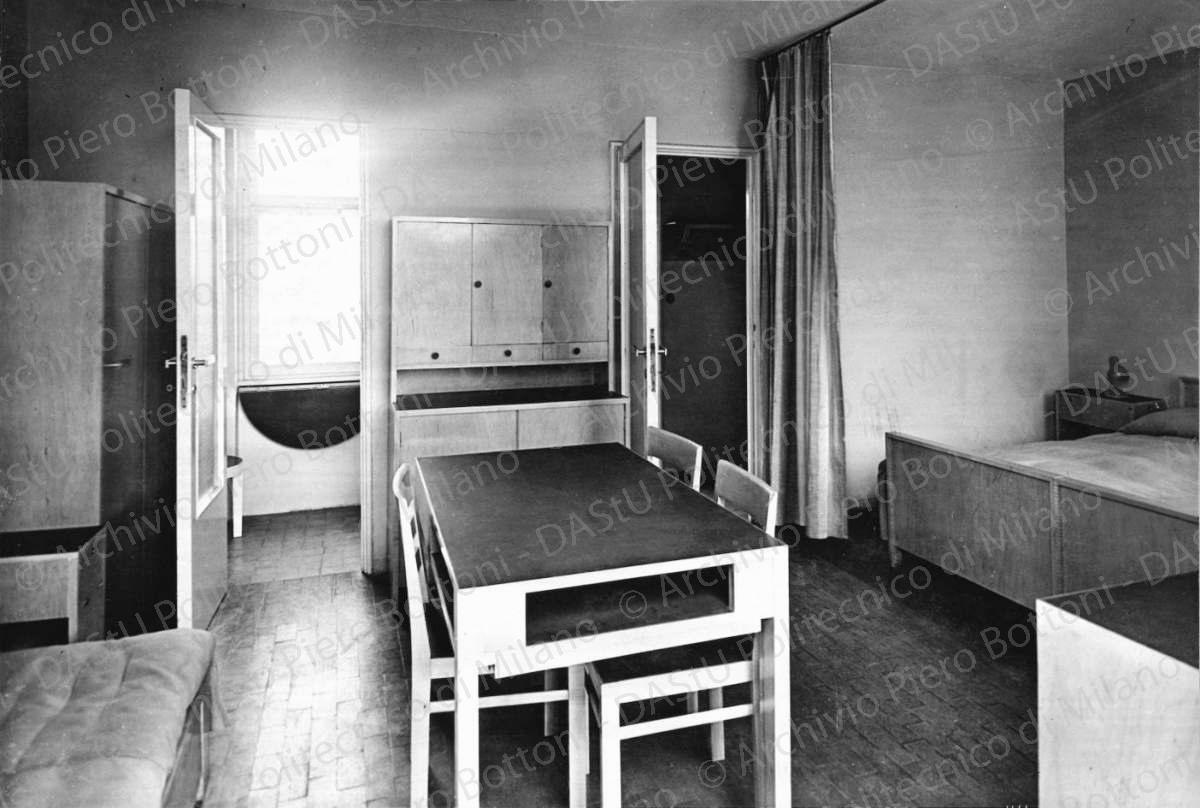 Veduta di un interno. Didascalia dattiloscritta: "1933 - Arredamento di alloggio popolare. La produzione di serie dell'arredo, applicata ad un tema sociale, partendo da elementi modulati con varianti nel dettaglio della finitura dei materiali poveri (legno o linoleum o laccatura) presentata alla V^ esposizione Triennale di Milano (1933) dove ancora imperavano le case in candoglia o i timpani e gli obelischi della 'Domus nova' (collaboratore ing. Griffini)". Positivo, bianco e nero, 201x297 mm, album giallo n. 19, pag.123.