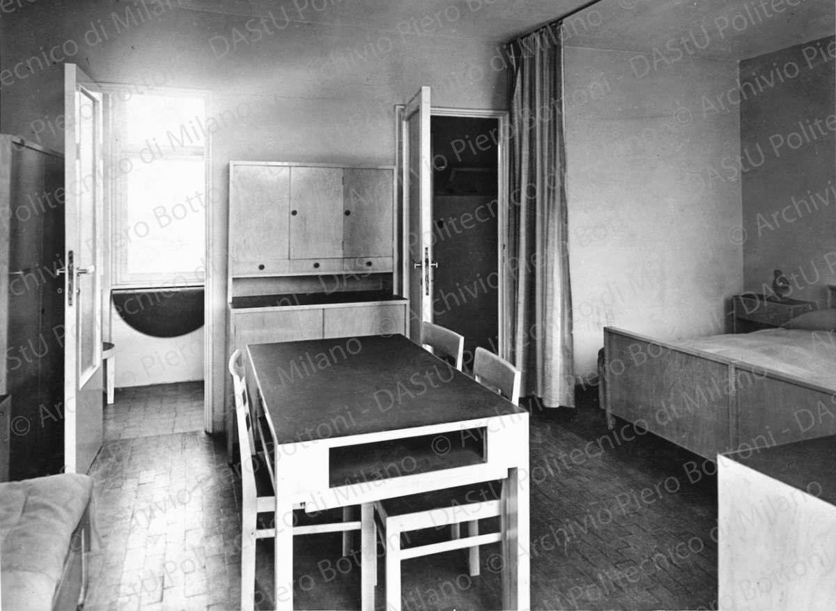Veduta di un interno. Didascalia: "1933. Arredamento di alloggio popolare. La produzione di serie dell'arredo, applicata ad un tema sociale, partendo da elementi modulati con varianti nei dettagli della finitura dei materiali poveri (legno o linoleum o laccatura) presentata alla V esposizione Triennale di Milano (1933) dove ancora imperavano le case in candoglia o i timpani e gli obelischi della 'Domus nova' (collaboratore ing. Griffini)". Positivo, bianco e nero, 202x275 mm, album giallo n. 7, pag. 29.