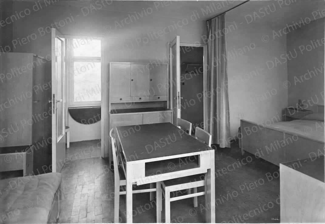 Veduta interna di un soggiorno. Didascalia: 'Alloggio n. 3 - mq 25,50; in fondo ingresso e W.C. e a sinistra cucina; in primo piano, a destra nicchia con tenda divisoria per letto matrimoniale (pavim. in linoleum); a sin. soggiorno con pavim. in piastrelle; mobili in ontano naturale e piani in linoleum (esecuz. Meroni e Fossati)'. Positivo, bianco e nero, 156x227 mm, album giallo n. 7, pag. 45.