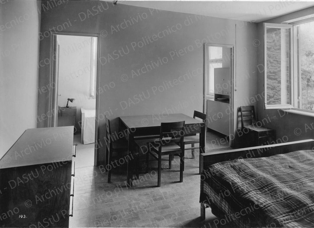 Veduta interna dell'alloggio n. 5. Didascalia: 'Locale di soggiorno e in fondo cucina e camera a 1 letto(mobili in insulite, piani in linoleum)'. Positivo, bianco e nero, 163x227 mm, album giallo n. 7, pag. 49.