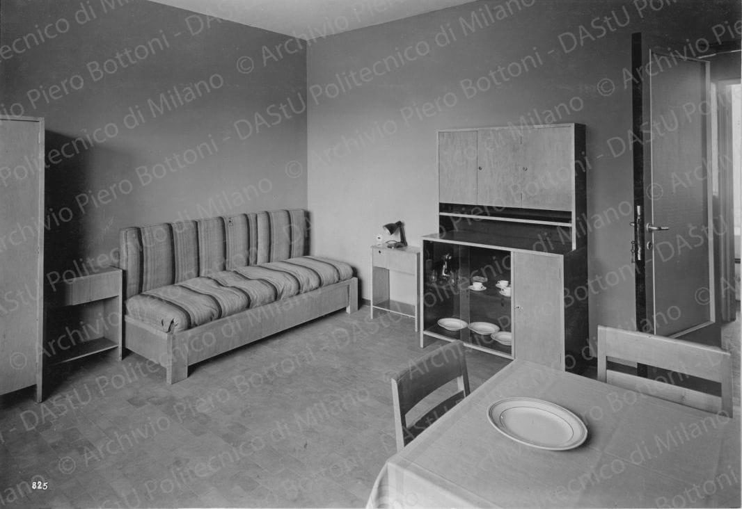 Veduta interna dell'alloggio n. 6. Didascalia: 'Cucina (a destra il passapiatti, mobili laccati piani in linoleum) e locale di soggiorno col divano-letto'. Positivo, bianco e nero, 156x226 mm, album giallo n. 7, pag. 51.