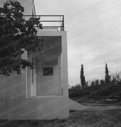 Veduta del soggiorno con il caminetto con libreria. Didascalia dattiloscritta: "1934 - Soggiorno nella casa di campagna a Reggio Emilia. Il tema compositivo è affidato ad alcuni semplicissimi elementi funzionali: il soppalco che taglia il salone, la libreria bassa, in muratura, che accoglie anche il caminetto, le appliques, la porta vetrata, chiara, la libreria ad essa normale, scura, elementi tutti composti nello spirito del razionalismo derivante dalla Bauhaus". Positivo, bianco e nero, 288x216 mm, album giallo n.19, pag.23.