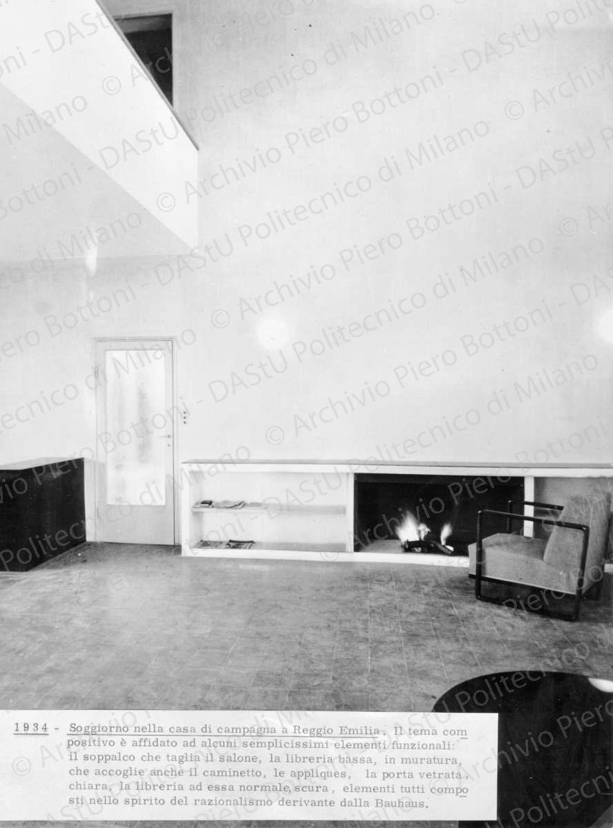 Veduta della "Libreria in muratura con caminetto", positivo, bianco e nero, mm 289x217. Annotazione dattiloscritta su carta incollata sul verso: "1934. Soggiorno nella casa di campagna a Reggio Emilia. Il tema compositivo è affidato ad alcuni semplicissimi elementi funzionali: il soppalco che taglia il salone, la libreria bassa, in muratura, che accoglie anche il caminetto, le appliques, la porta vetrata, chiara, la libreria ad essa normale, scura, elementi tutti composti nello spirito del razionalismo derivante dalla Bauhaus".
