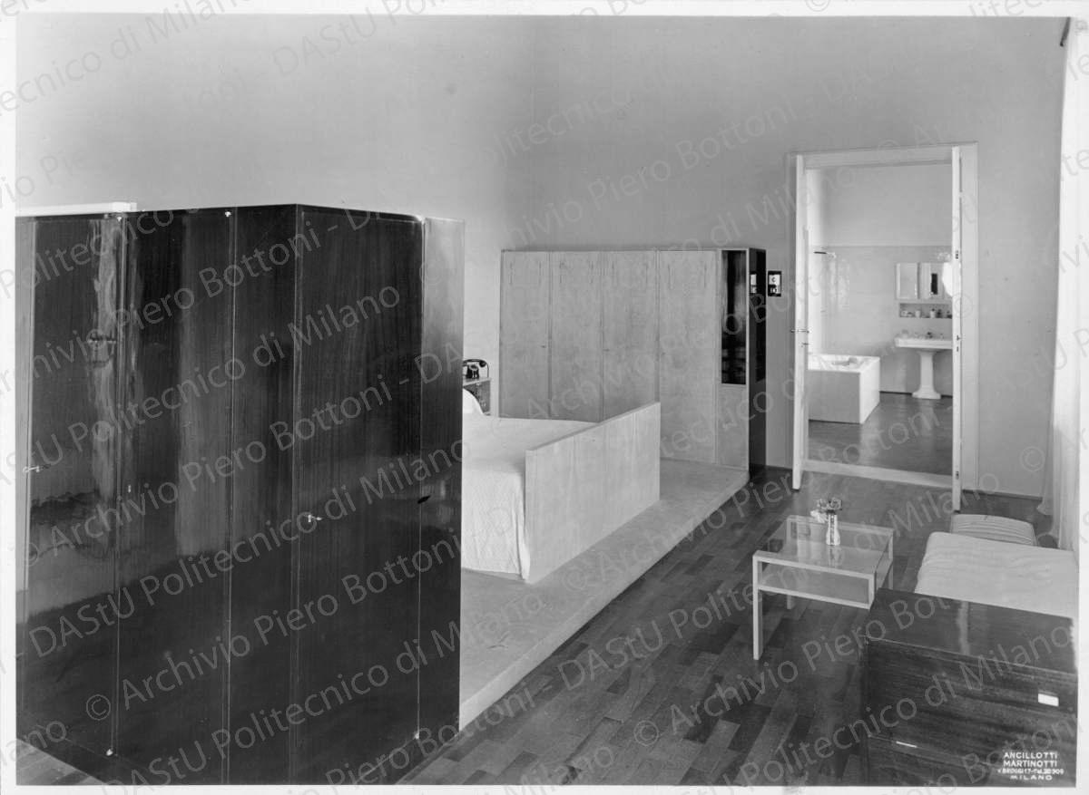 Camera da letto dei genitori, positivo, bianco e nero, mm 170x232. Annotazione dattiloscritta su velina incollata sul verso della fotografia: "La camera da letto dei genitori è divisa in due zone mediante una parete armadio. Nella prima zona adibita a spogliatoio gli armadi e la toeletta sono in legno di palissandro, mentre nell'altra il letto in legno di acero chiaro, con coperta a colori vivaci, è fiancheggiato da pareti-armadio pure in acero. Una pedana ricoperta in fustagno verde chiaro distingue la zona riservata per la notte dalla zona di passaggio in cui sono disposti un divano ricoperto in fustagno verde ed un tavolino di acero e cristallo. La camera comunica col bagno ed i servizi rivestiti in opalina verde chiara a grandi lastre".